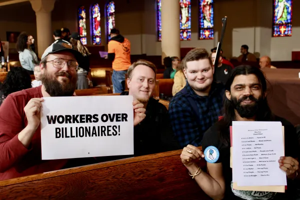 Group of men smiling and sitting together in a church pew holding a sign that says “Workers Over Billionaires”