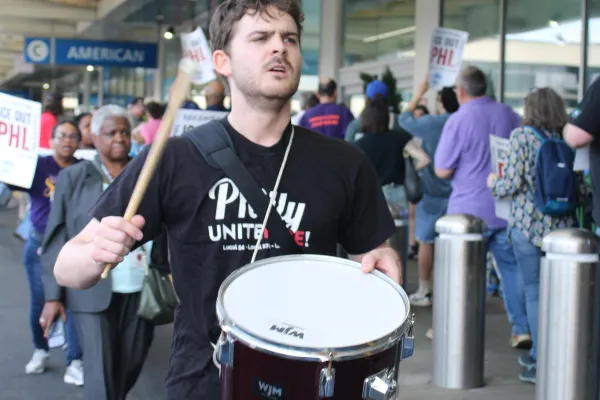 A young man wearing a black “Philly Unite Here” t-shirt walking a picket line while playing a snare drum.