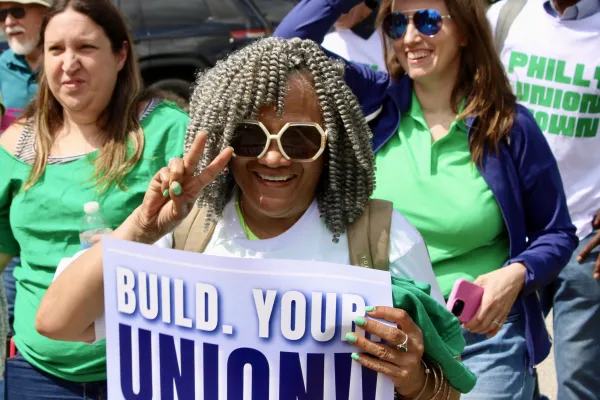 A woman smiling widely at the camera while walking up the street holding a sign that says “Build Your Union!”