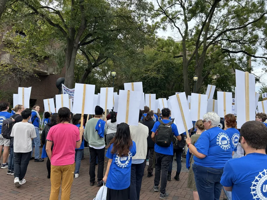 A group of grad students with their backs to the camera and wearing blue UAW shirts while holding picket signs
